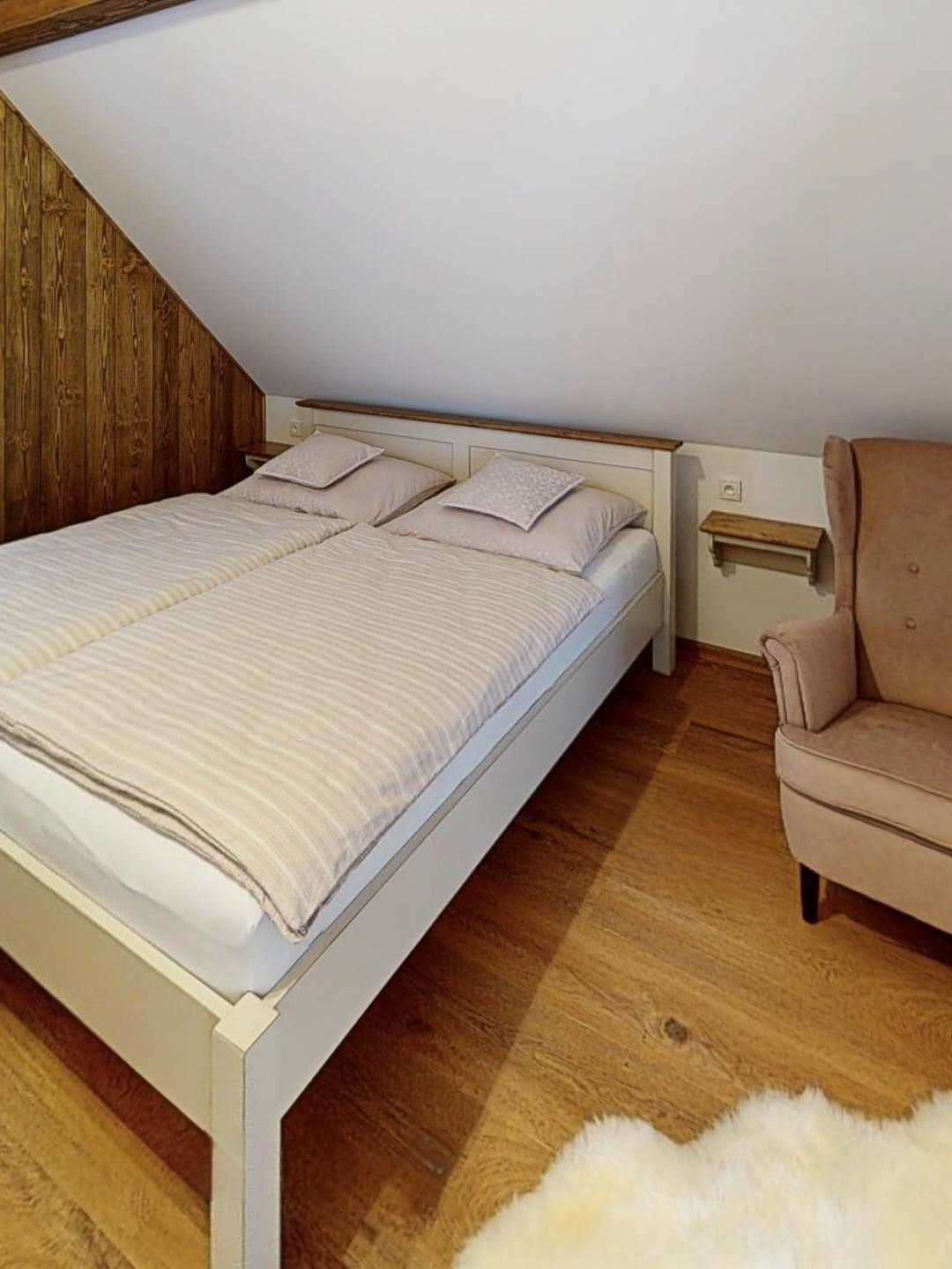 Ložnice pod šikmou střechou s pohodlnou manželskou postelí, světlým dřevem a relaxačním křeslem.