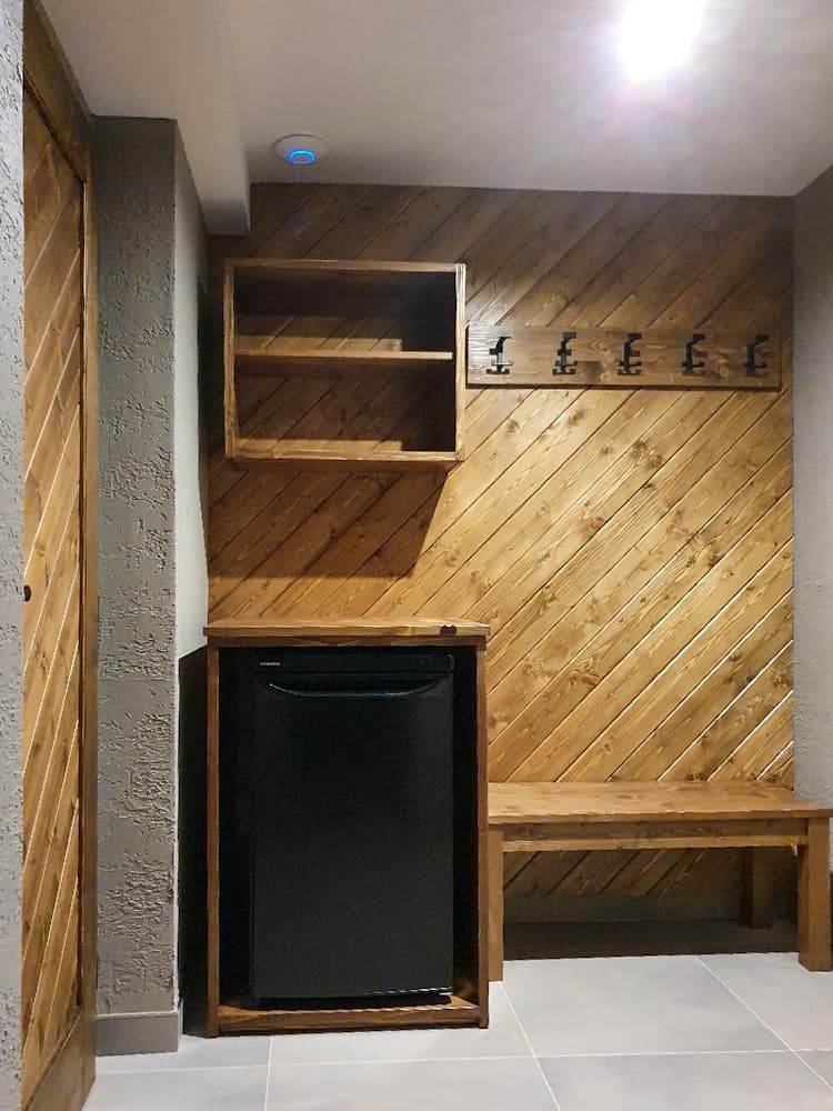Zázemí s malou chladničkou, dřevěnou stěnou, úložnou skříňkou a lavicí v suterénu apartmánu.