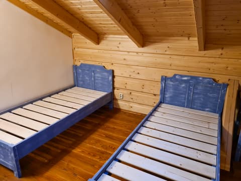 Dvojice masivních dřevěných postelí s modrým rámem a laťkovými rošty uspořádaná v dřevěném podkroví selského stavení.